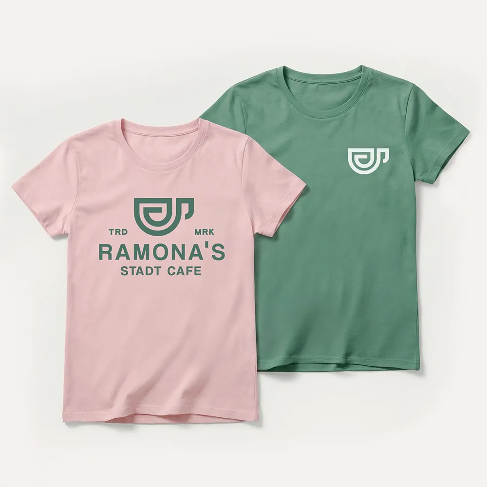 Zwei T-Shirts nebeneinander, eines rosa mit grünem Logo und dem Text „Ramona’s Stadt Cafe“ und das andere grün mit kleinem weißem Logo.