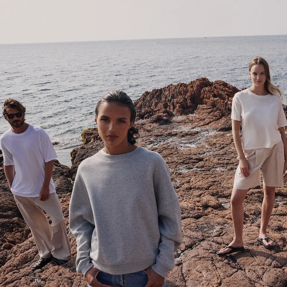 Drei Personen stehen auf felsigem Gelände am Meer und tragen lässige, minimalistische Kleidung in neutralen Farbtönen.
