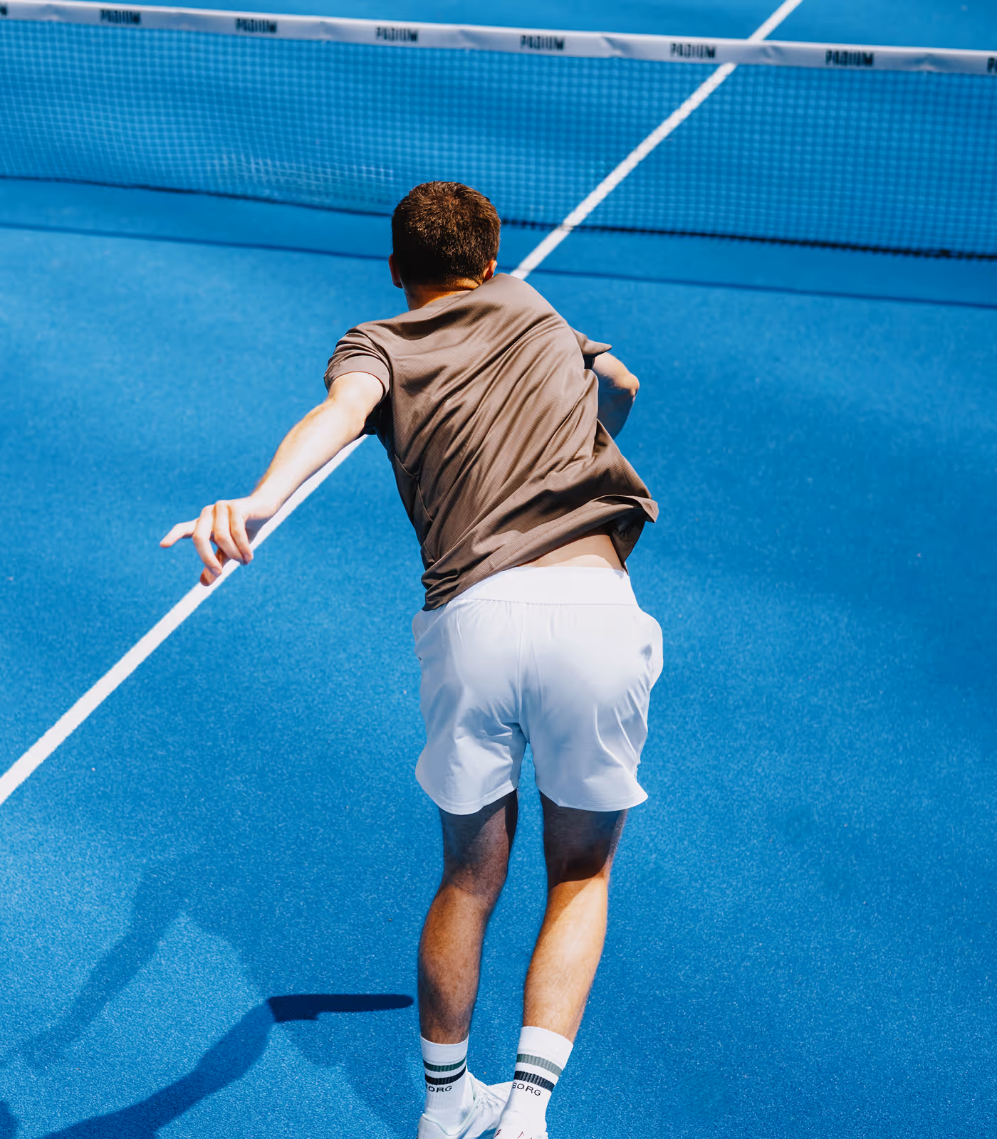 Man in white shorts and brown shirt playing padel on a blue court with a padel racket in his right hand.
