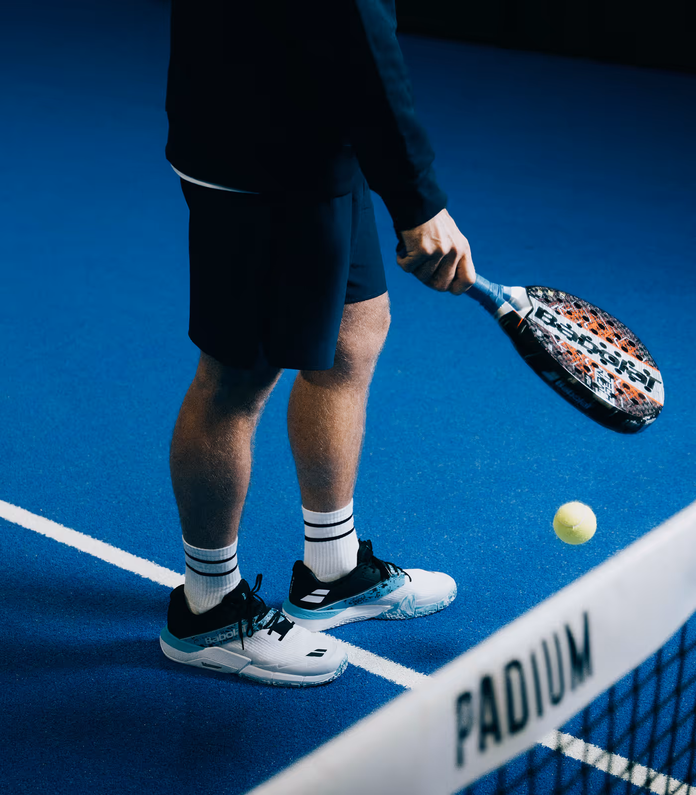 Player on blue padel court preparing to hit yellow ball with Babolat paddle near net labeled PADIUM.