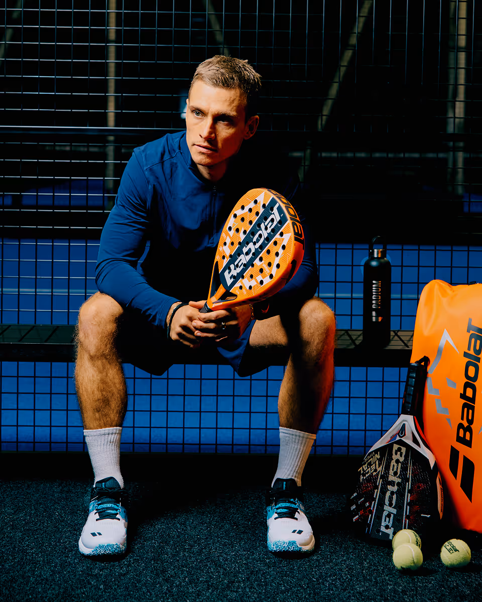 Athlete sitting on a bench holding an orange Babolat padel racket with tennis balls, extra racket, water bottle, and Babolat bag nearby.