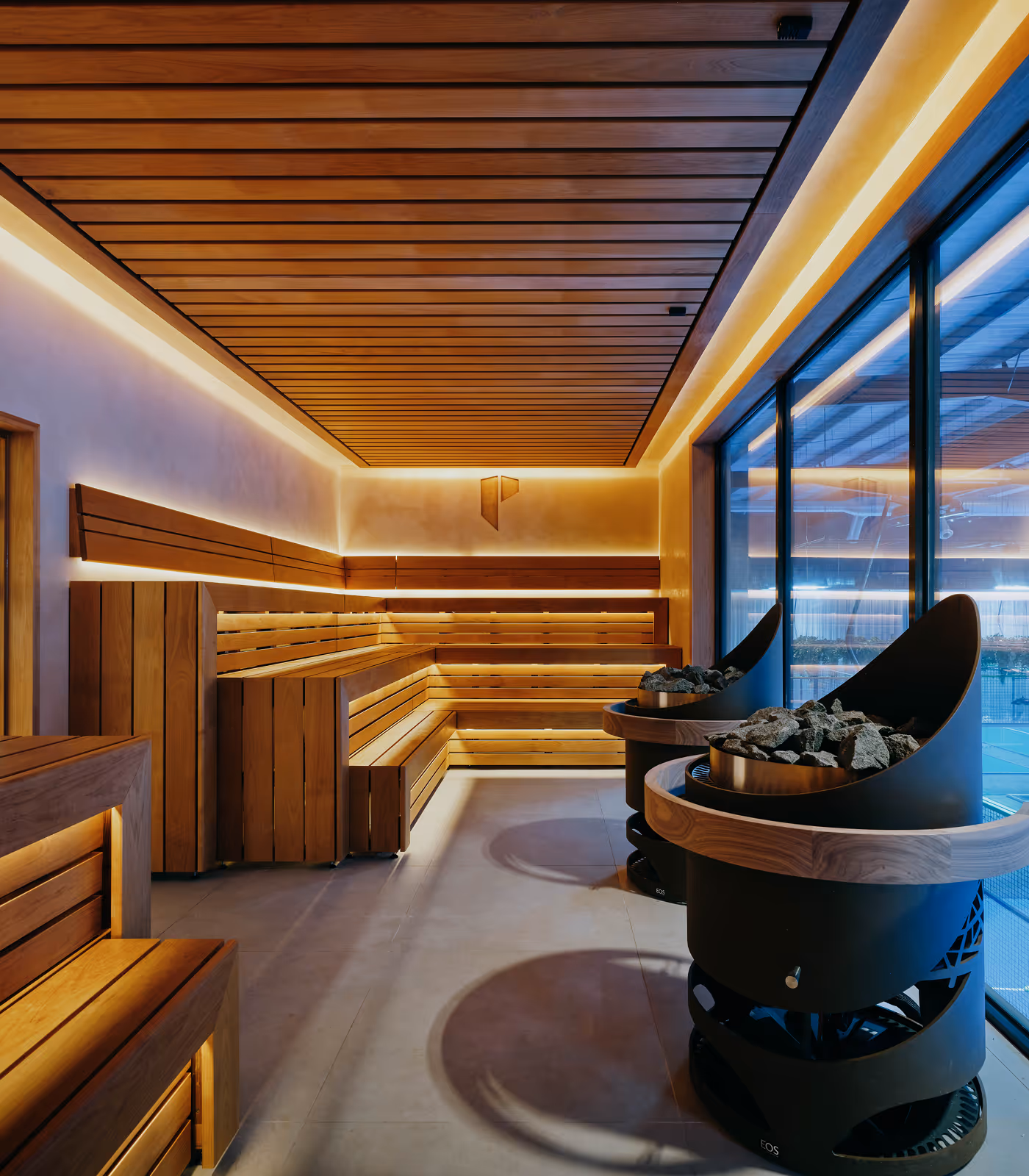 Modern sauna interior with wooden benches, ceiling panels, ambient lighting, and two black sauna heaters filled with stones near large windows.