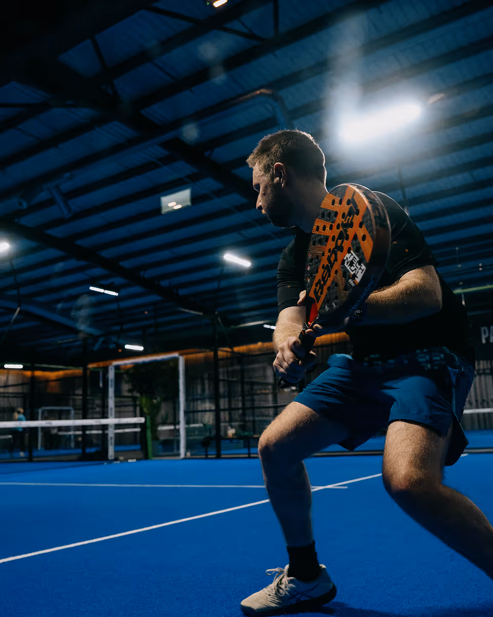 Man in blue shorts playing padel indoors holding a paddle racket in a ready stance.