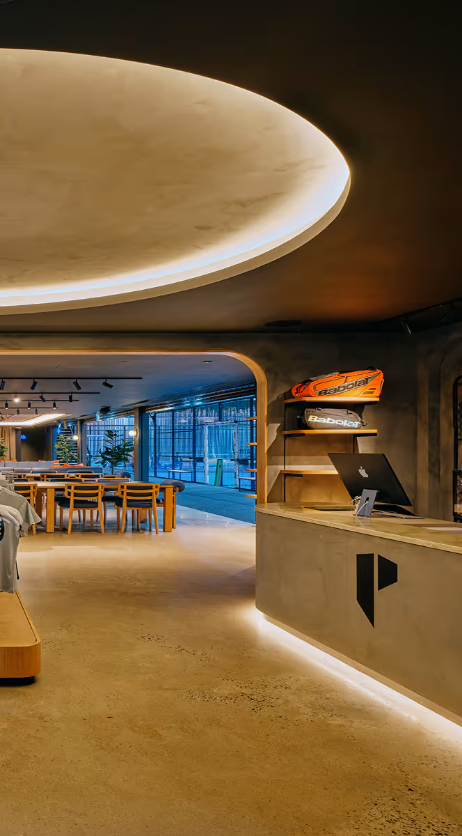 Modern sports facility interior with a reception desk, wooden tables and chairs, and tennis racket bags displayed on shelves.