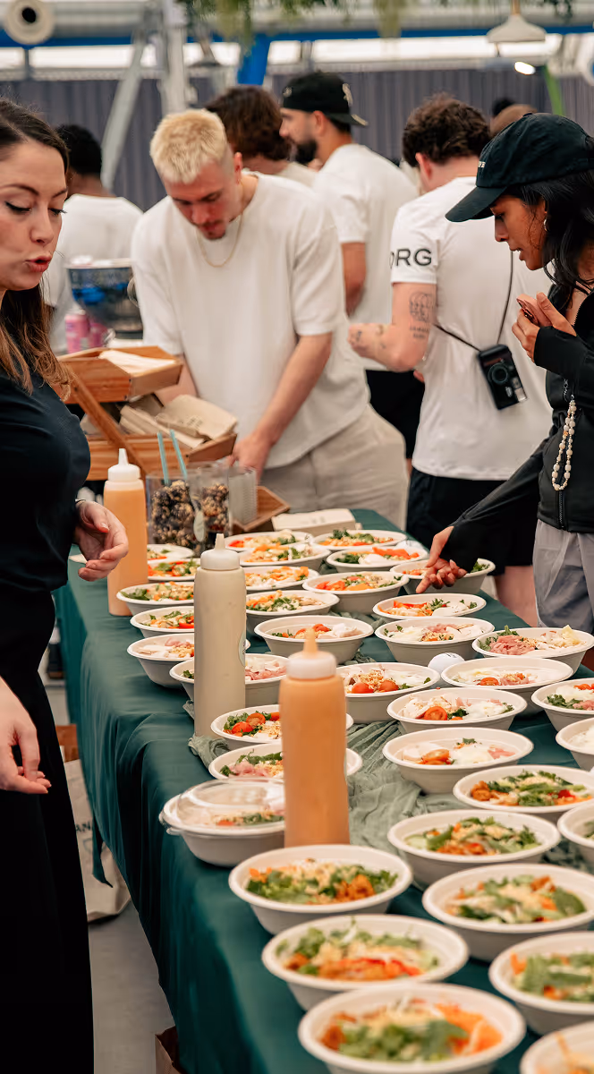 People serving and selecting multiple bowls of fresh salads on a table with sauce bottles during an event.