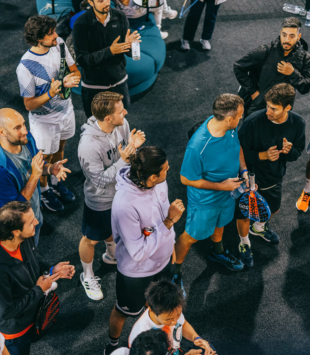 Group of men standing on a tennis court holding paddle rackets and clapping, some wearing sportswear.