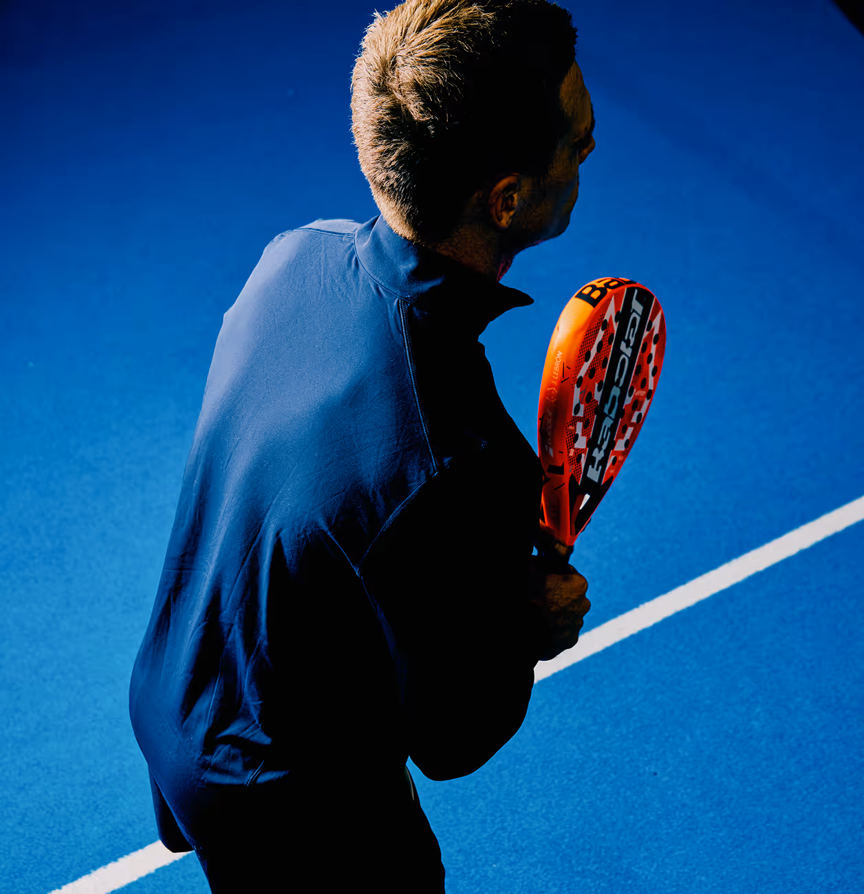 Man in a dark blue shirt holding an orange padel racket on a blue padel court.