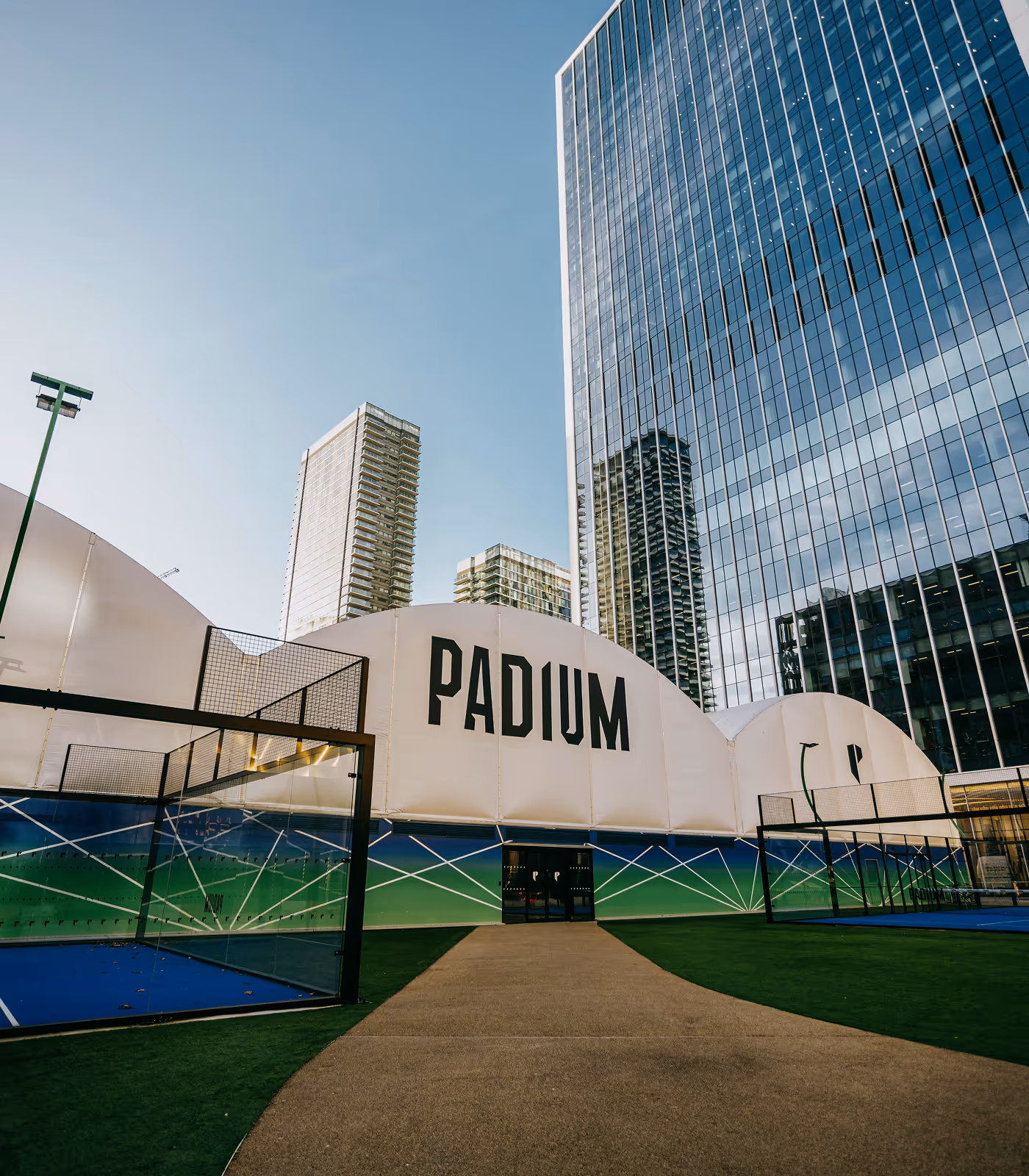 Outdoor sports courts with a large white tent structure labeled 'PADIUM' in front of modern glass skyscrapers under a clear blue sky.