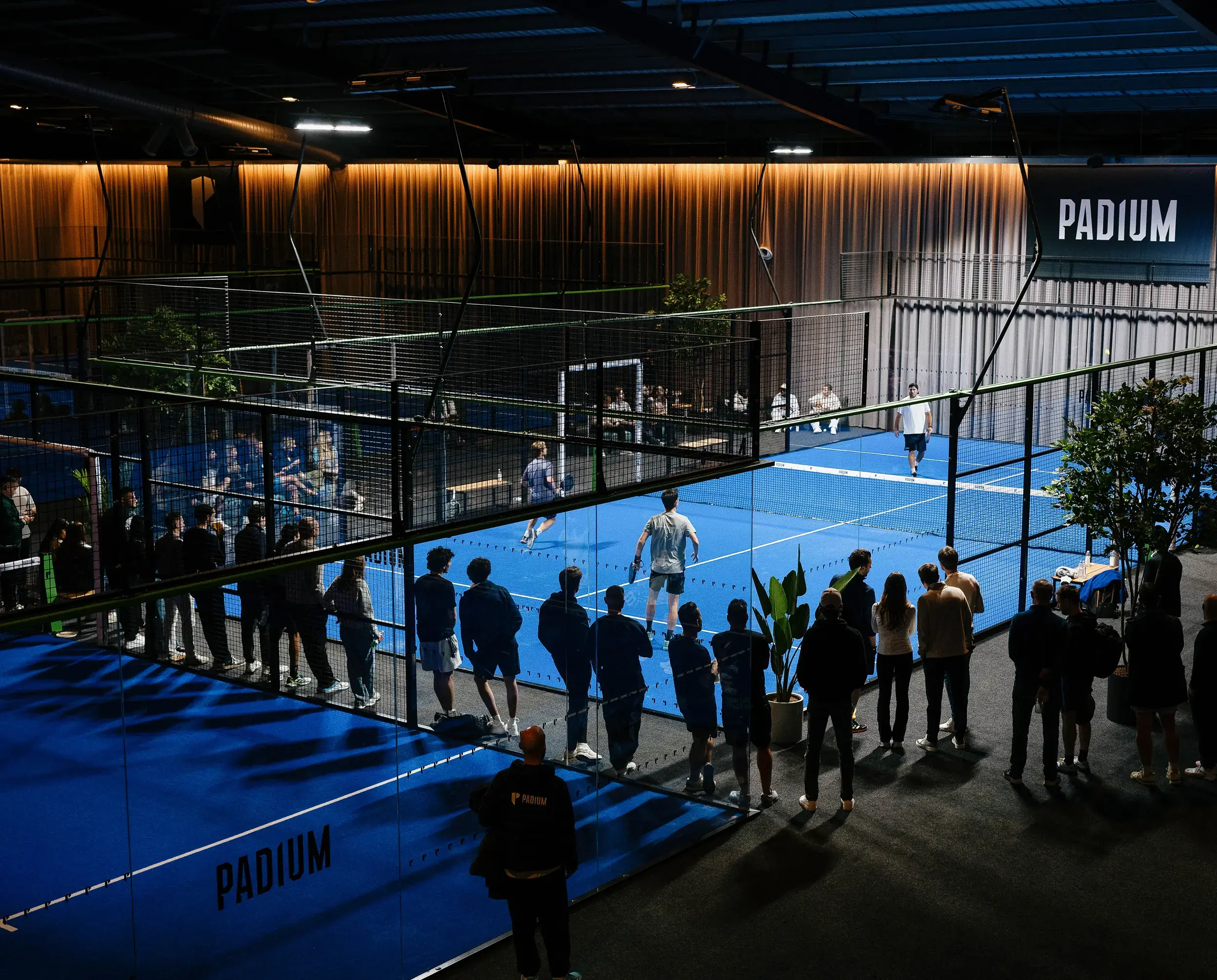 Indoor padel court with players in action and spectators watching from outside the fenced area.