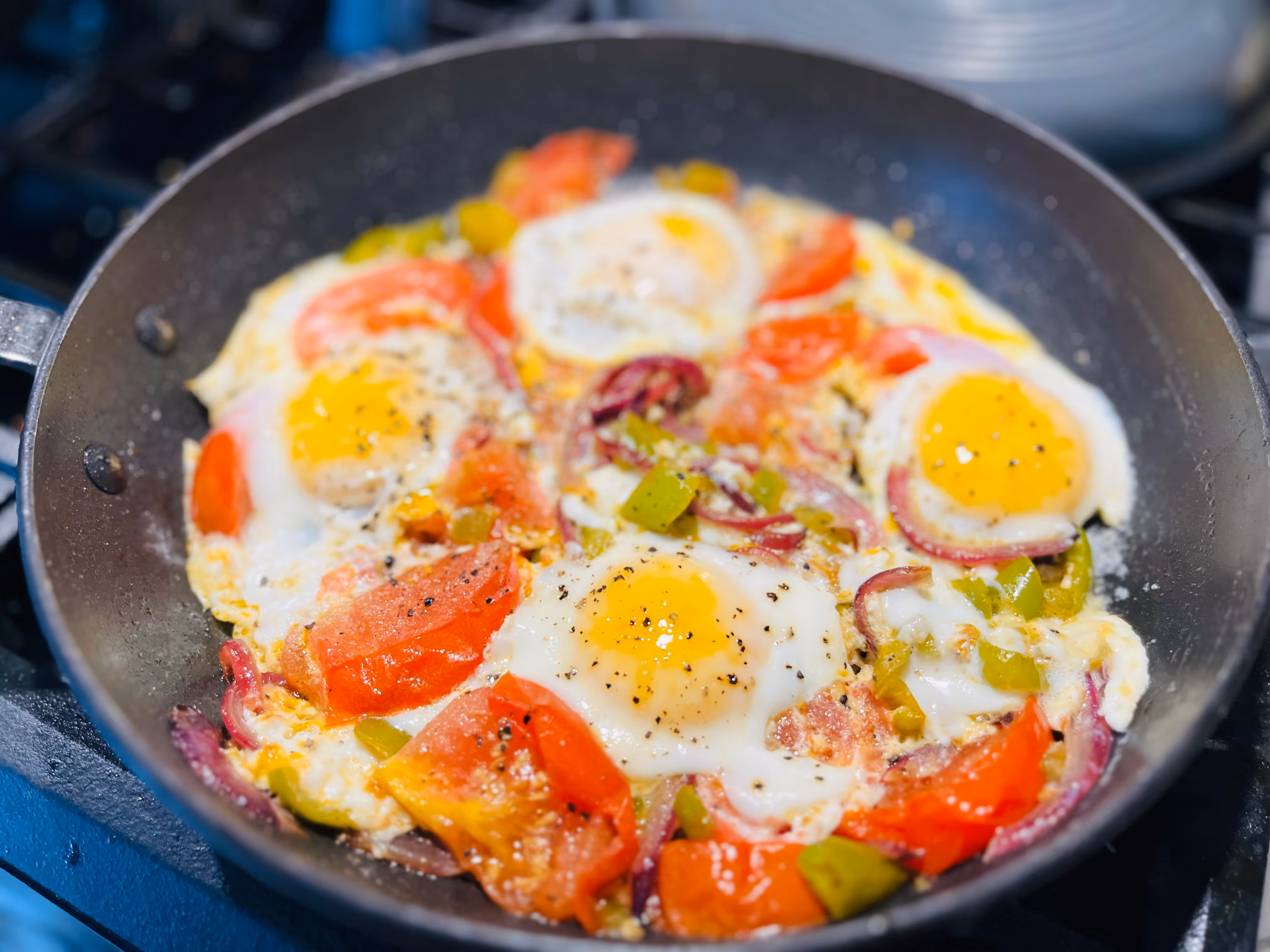 Four over easy eggs, tomatoes, and peppers cooking in a skillet