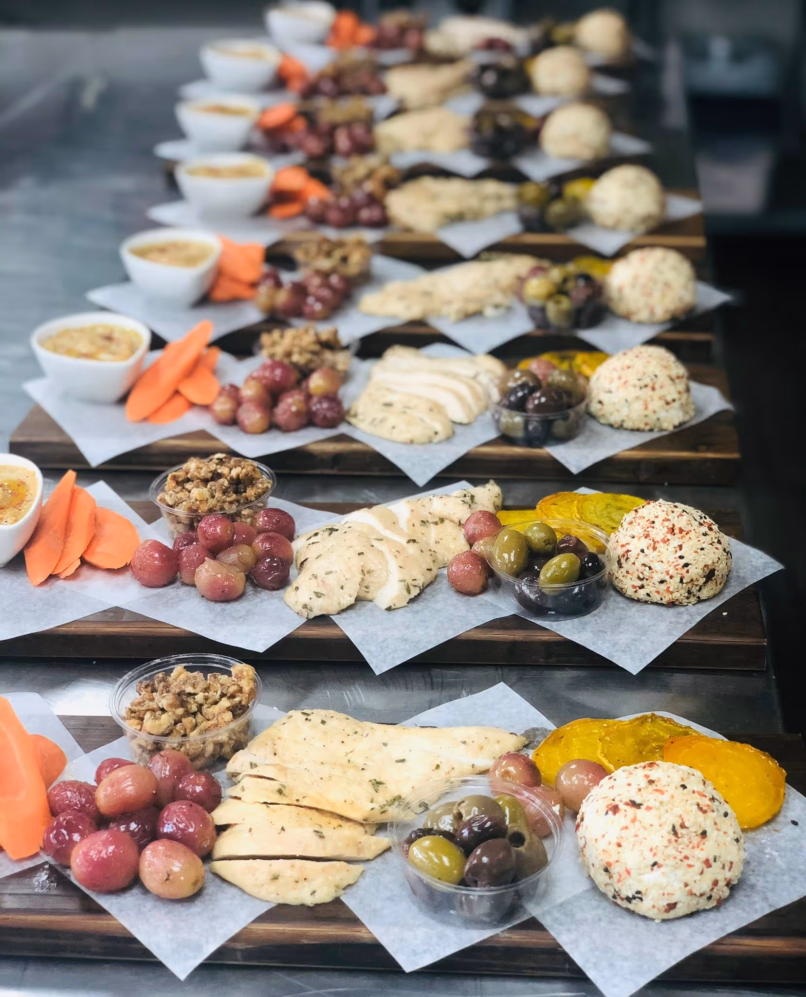 A series of rectangular wooden boards arranged with sliced meats, assorted nuts, and olives, paired with hummus, fresh pita, and seasonal roasted fruits."