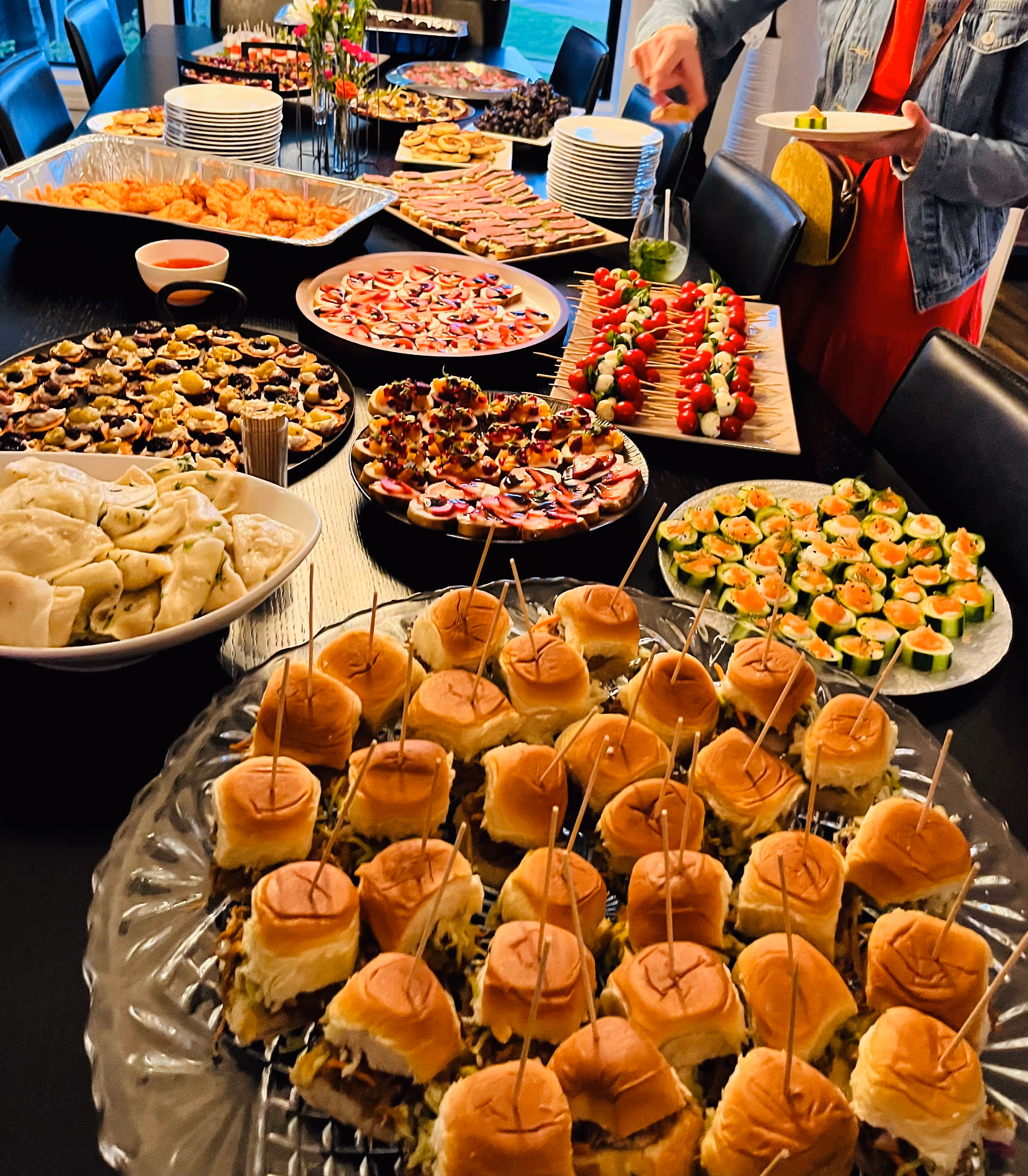 A wide perspective of an expansive catering spread featuring over a dozen platters, including gourmet mini sliders and a diverse array of colorful small bites.
