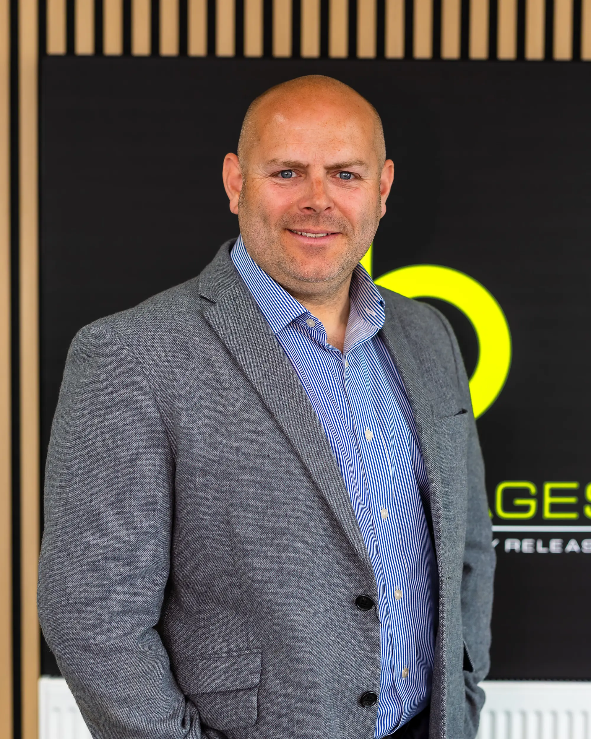 Smiling bald man in a gray blazer and striped blue shirt standing indoors with a black and yellow background.