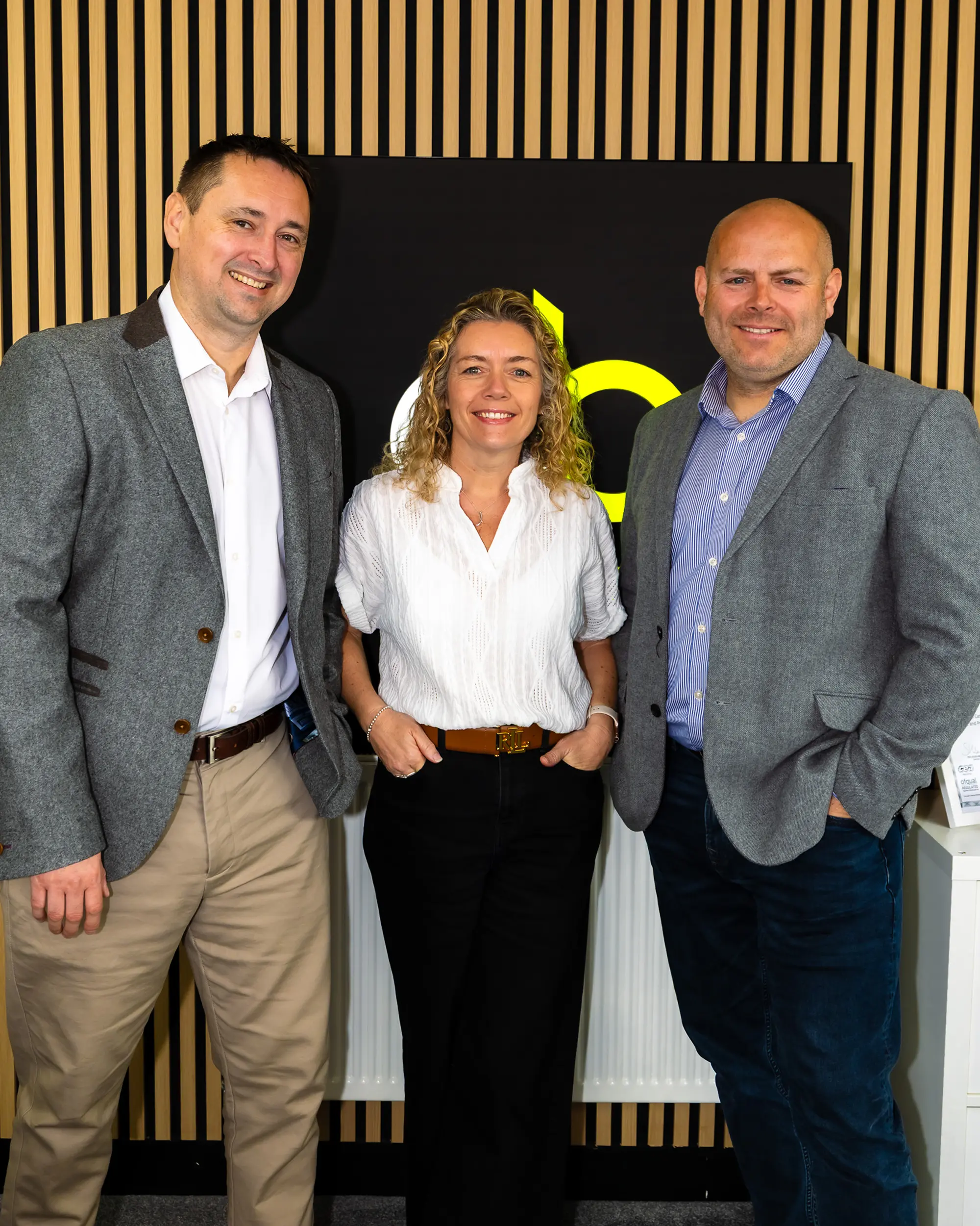 Three business professionals smiling, standing before a black and yellow company logo on wood panel wall.