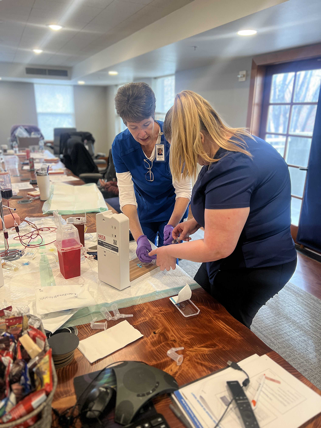 Kari and a healthcare professional student in blue scrubs practicing medical procedures together on a wooden table in a well-lit room.