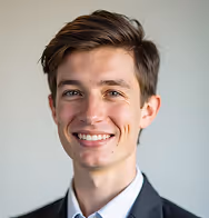 Smiling young man with short brown hair wearing a dark blazer over a light blue shirt against a neutral background.