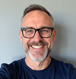 Smiling middle-aged man with short gray hair, beard, and glasses wearing a navy blue shirt against a gray background.