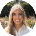 Smiling woman with long blonde hair wearing a white shirt, outdoors with blurred greenery and flowers in the background.