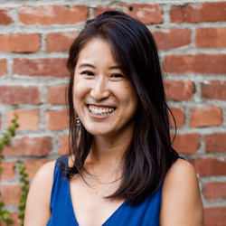 Smiling woman with long dark hair wearing a blue sleeveless top standing in front of a brick wall.