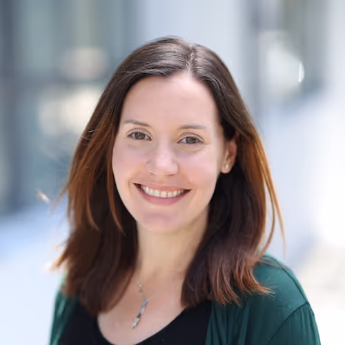 Smiling woman with medium-length brown hair wearing a dark green cardigan and black top against a blurred outdoor background.