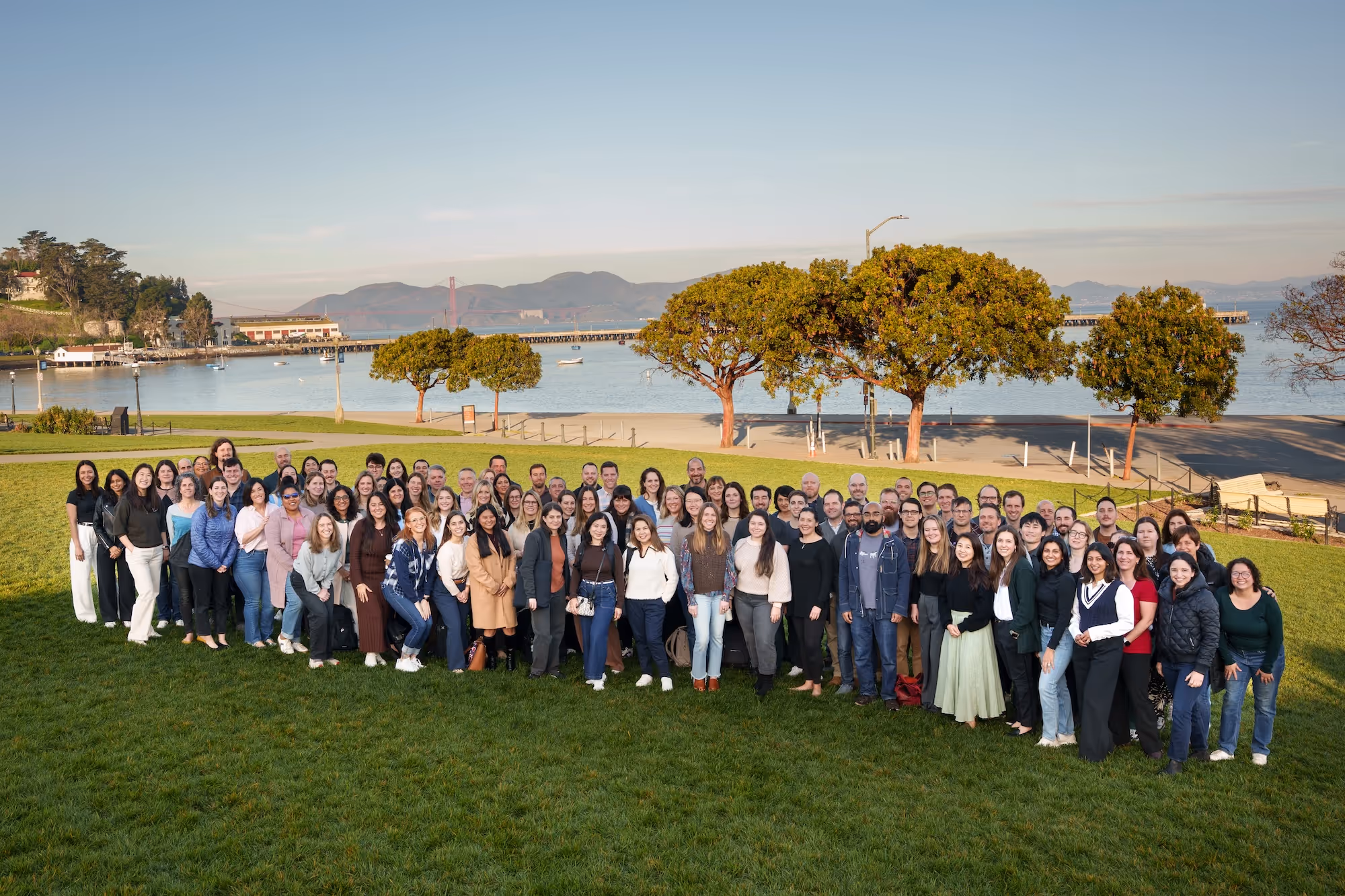 Large group of people standing on a grassy area with trees and a body of water, with the Golden Gate Bridge and hills in the background.