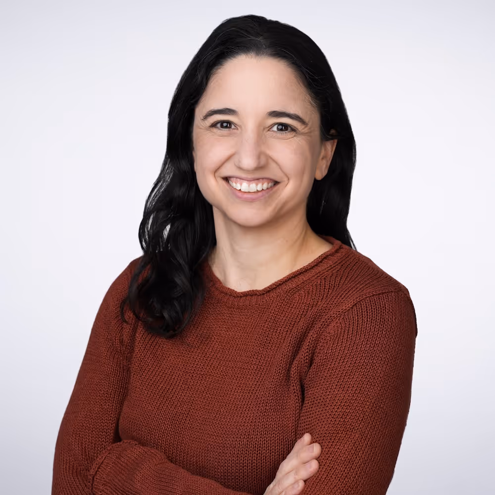 Smiling woman with dark hair wearing a rust-colored knitted sweater against a plain light background.