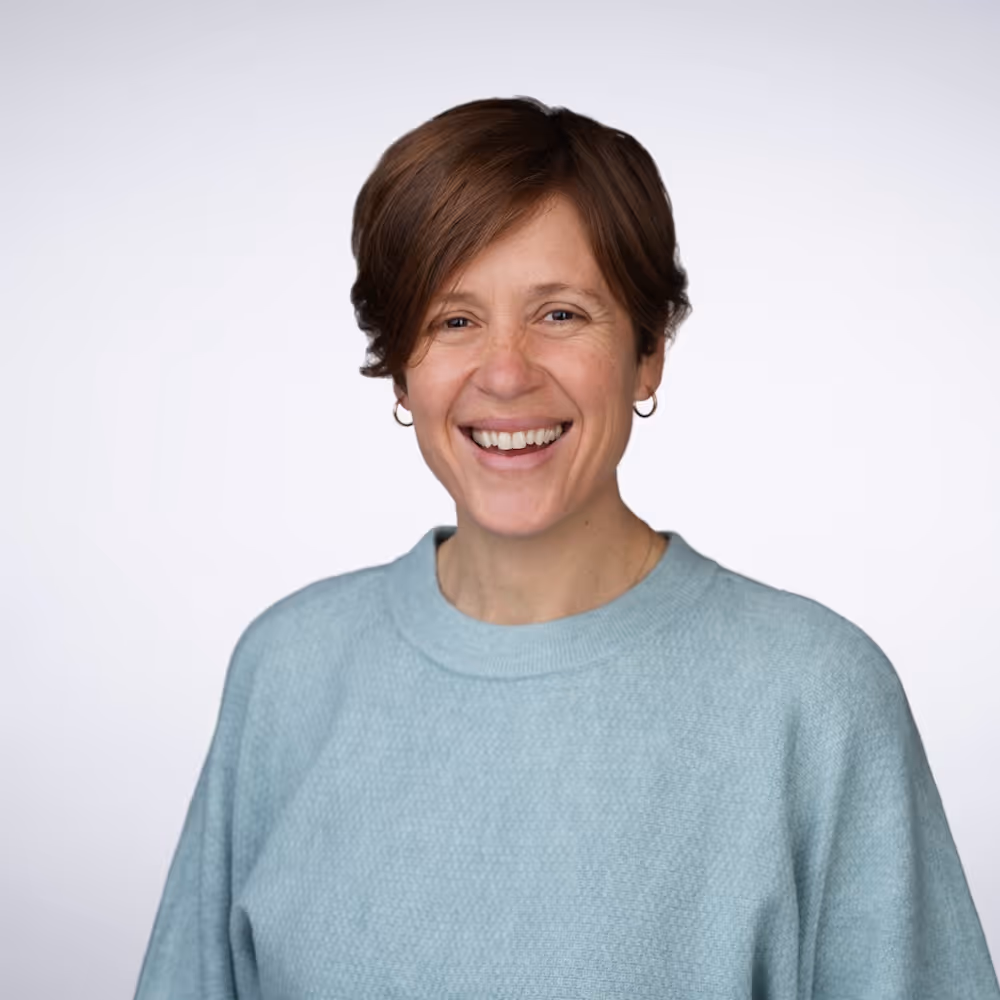 Smiling woman with short brown hair wearing a light blue sweater and hoop earrings against a white background.
