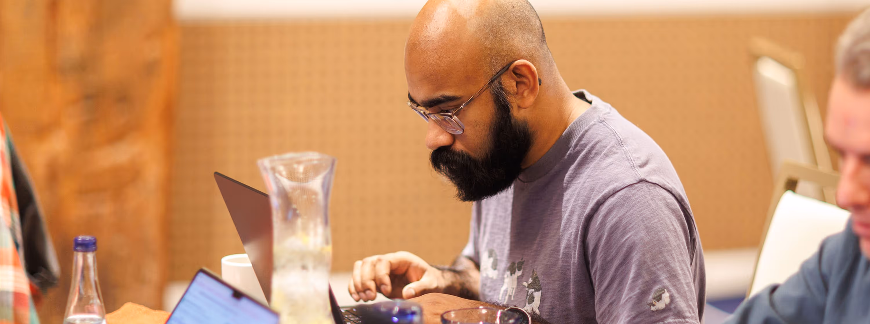 Man with glasses and beard working intently on a laptop at a table with a water pitcher and bottle.