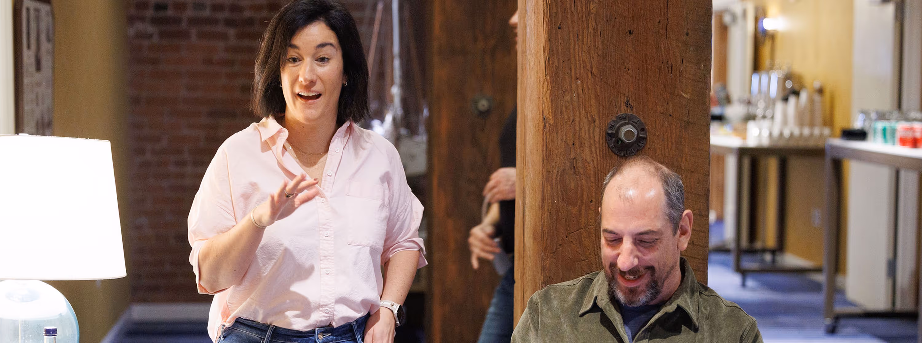 Woman in a pink shirt standing and gesturing with a man sitting and smiling beside a wooden pillar in a casual office setting.