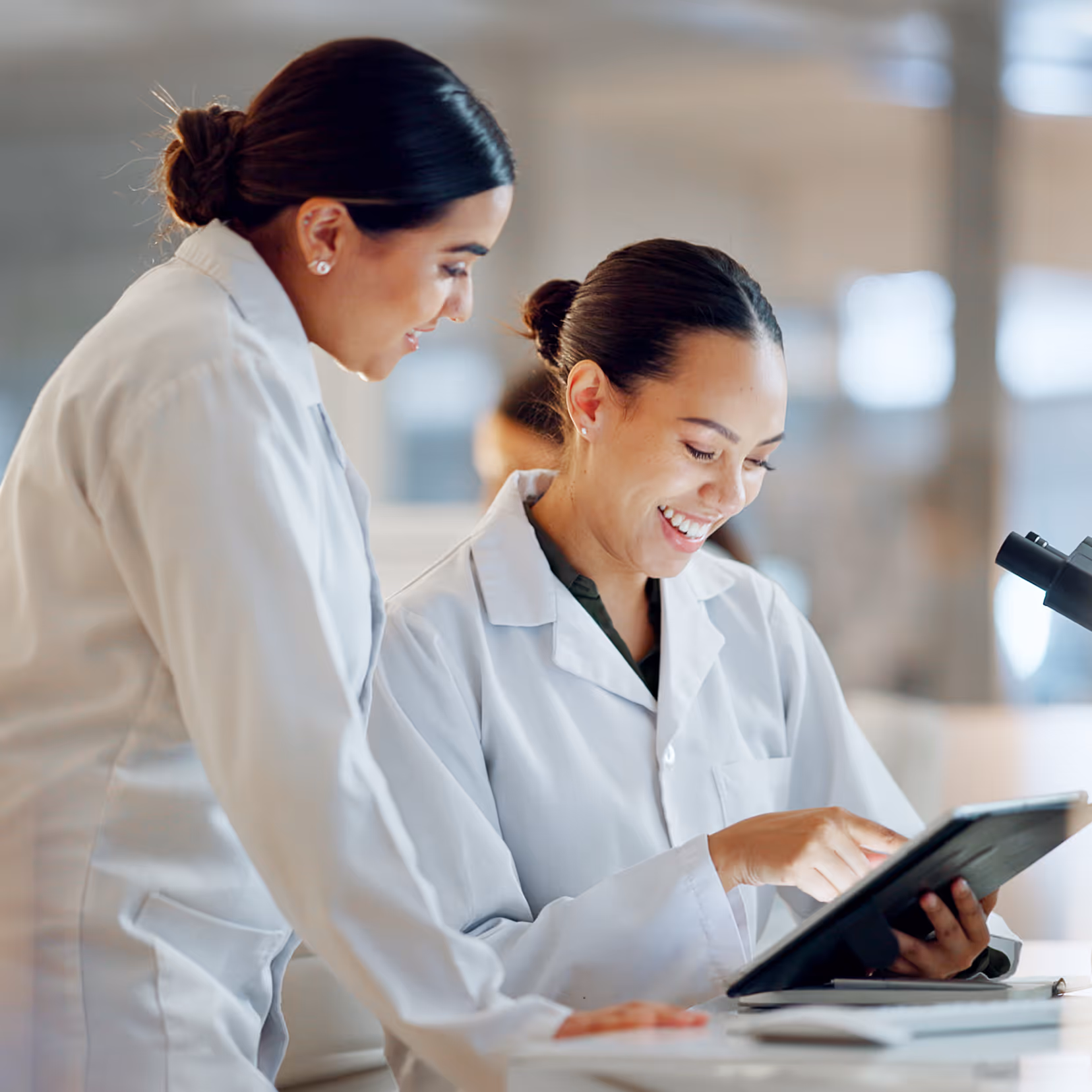 Two female scientists in lab coats smiling and using a tablet together in a laboratory.