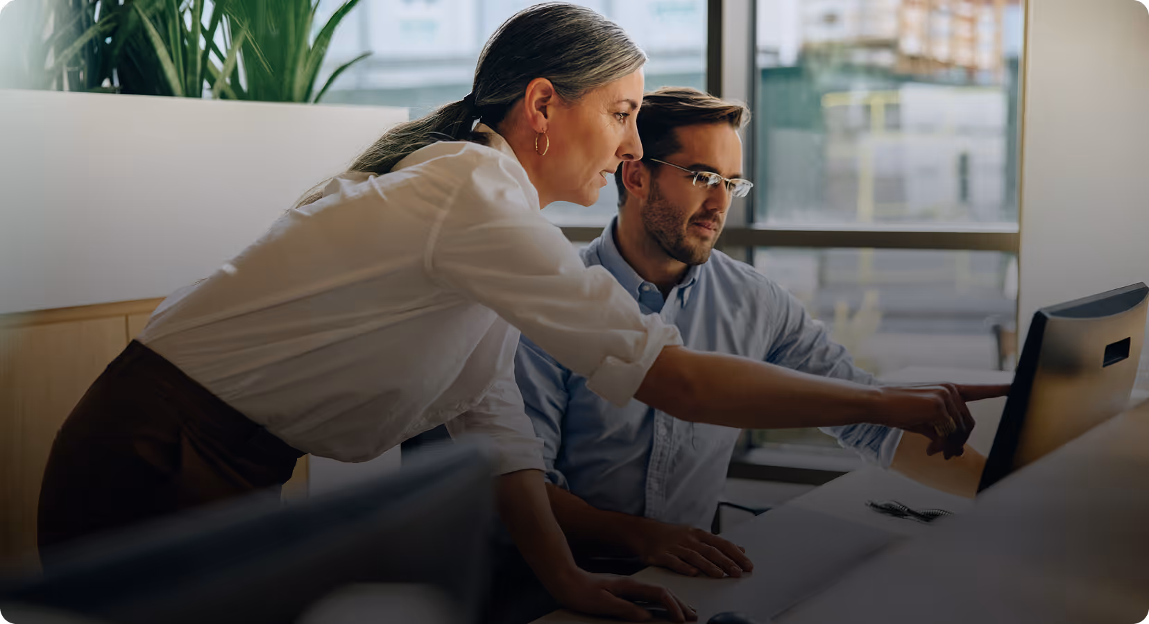 Woman in white shirt points at computer screen while man in blue shirt watches attentively in office.