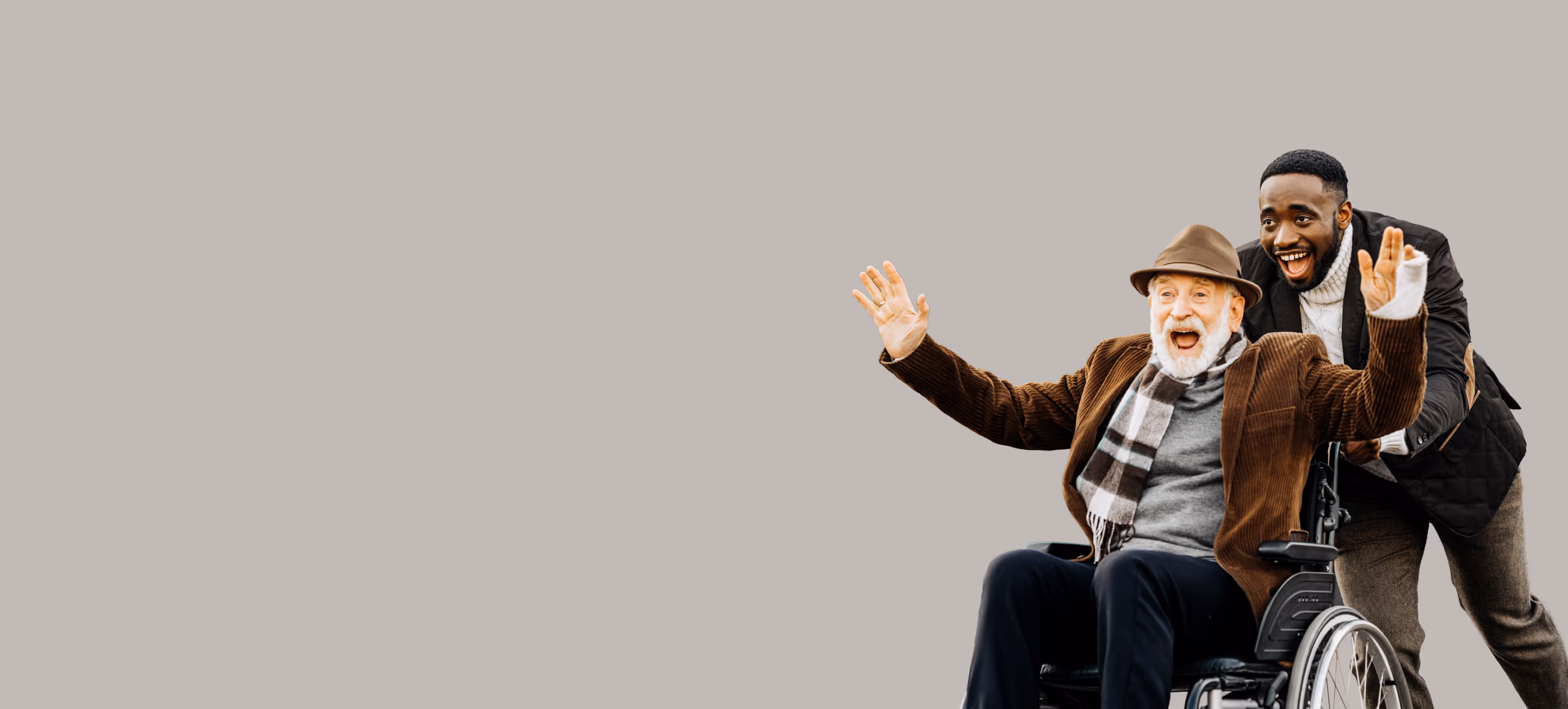 Smiling young man pushes happy elderly man with beard in wheelchair, both waving hands.