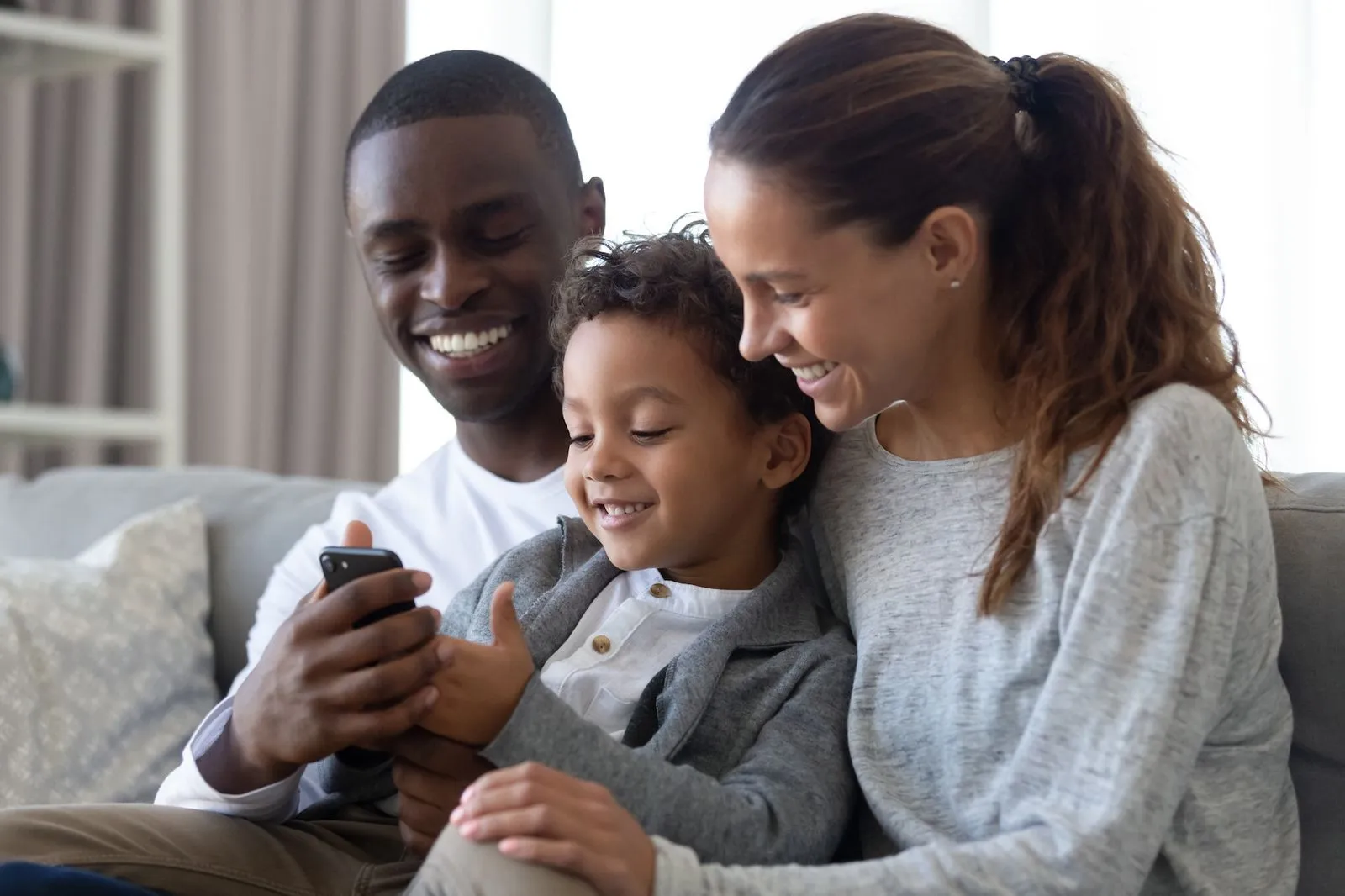 Smiling family of three sitting on a couch, looking at a smartphone together.