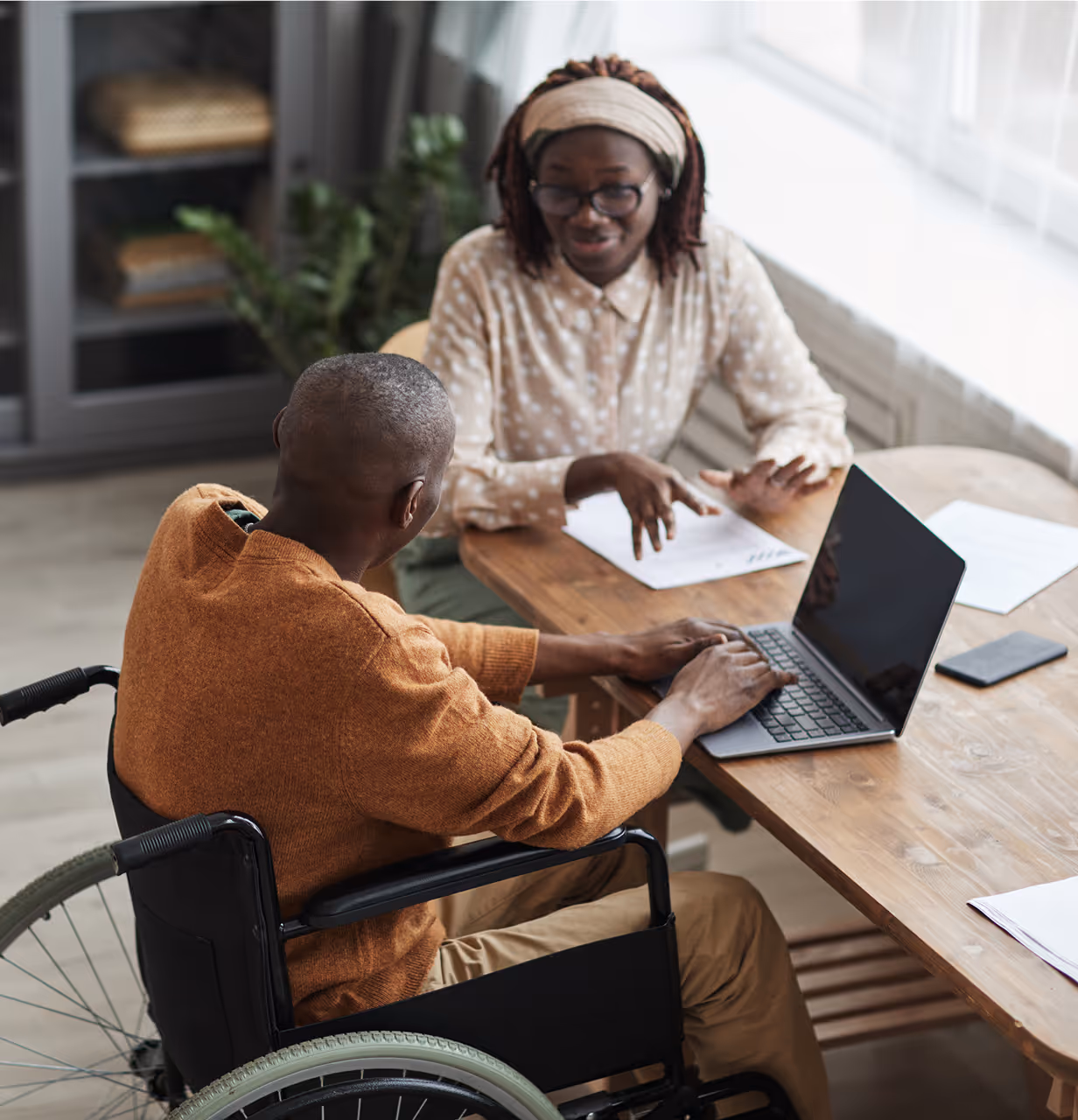 Man in a wheelchair using a laptop while a woman in glasses explains something across the table.