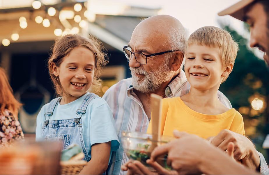Smiling elderly man with glasses and two happy children enjoying a meal outdoors together.