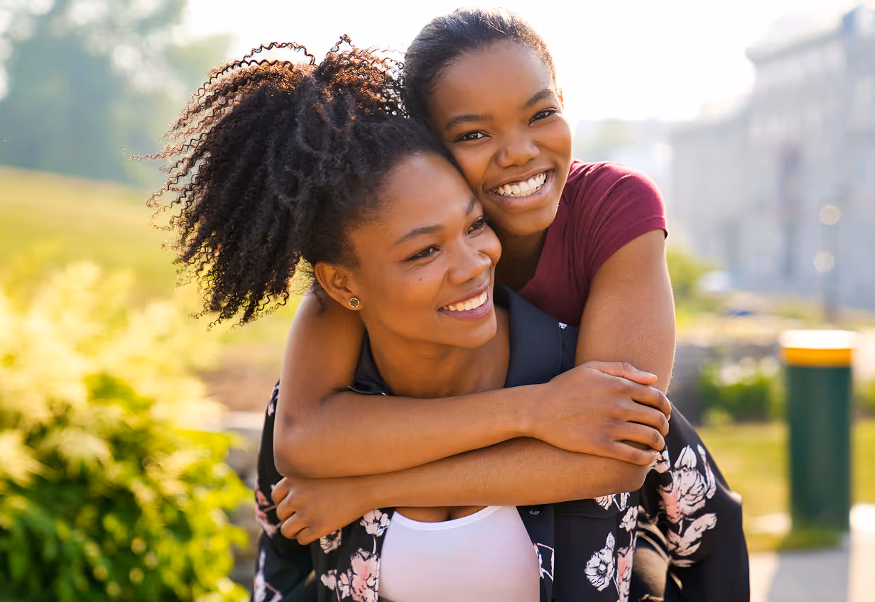 Smiling young woman giving a piggyback ride to a teenage girl outdoors on a sunny day.