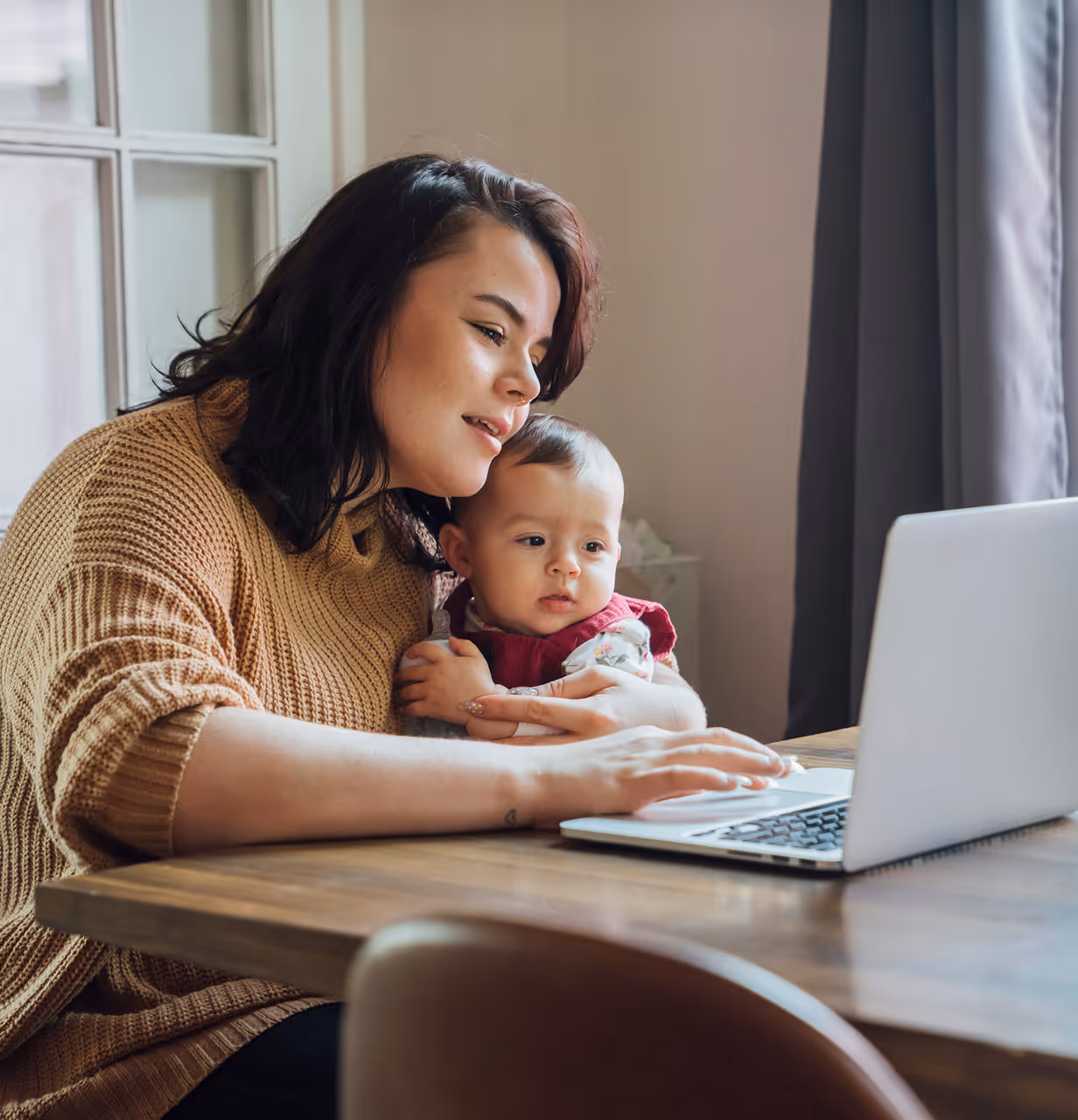 Smiling man in an orange shirt working on a laptop at a wooden desk with an open notebook and smartphone.