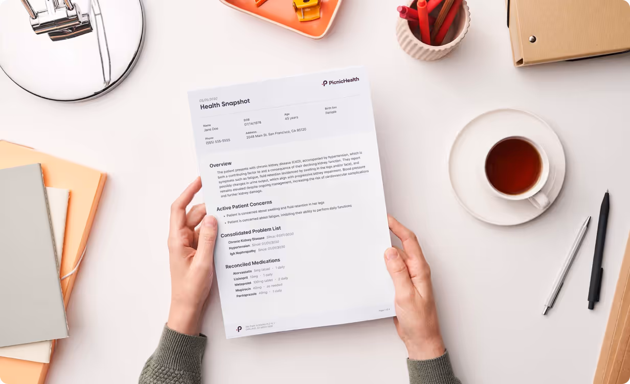 Hands holding a printed health snapshot report on a desk with notebooks, pens, a cup of tea, and an orange container with stationery.