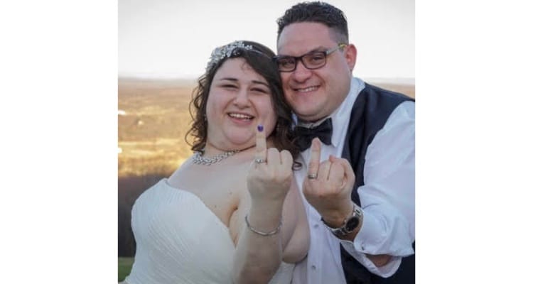 Two people in wedding outfits face the camera with their ring fingers raised. They’re both smiling.