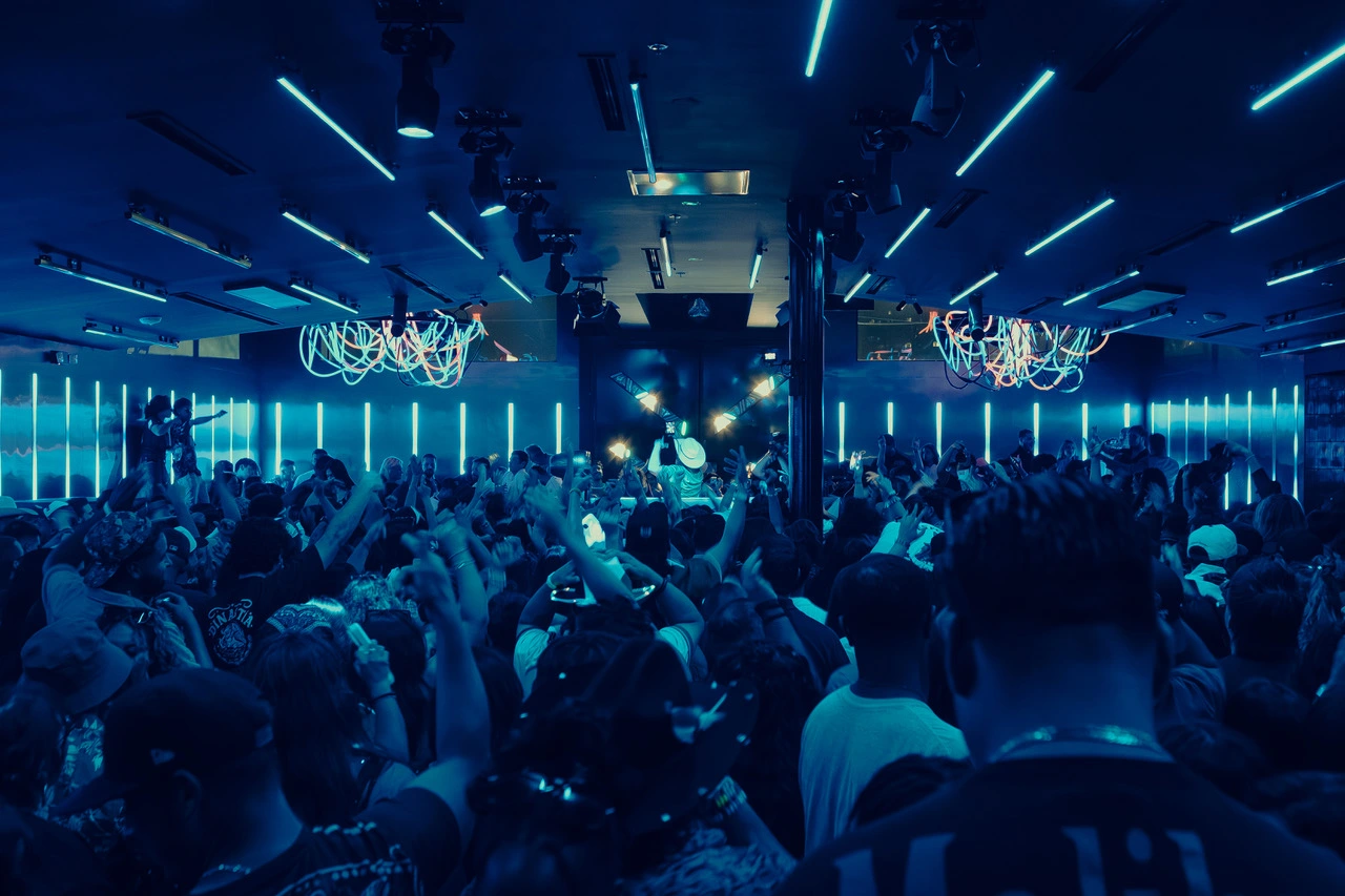 Crowded indoor concert with blue neon lighting and a DJ performing on stage.