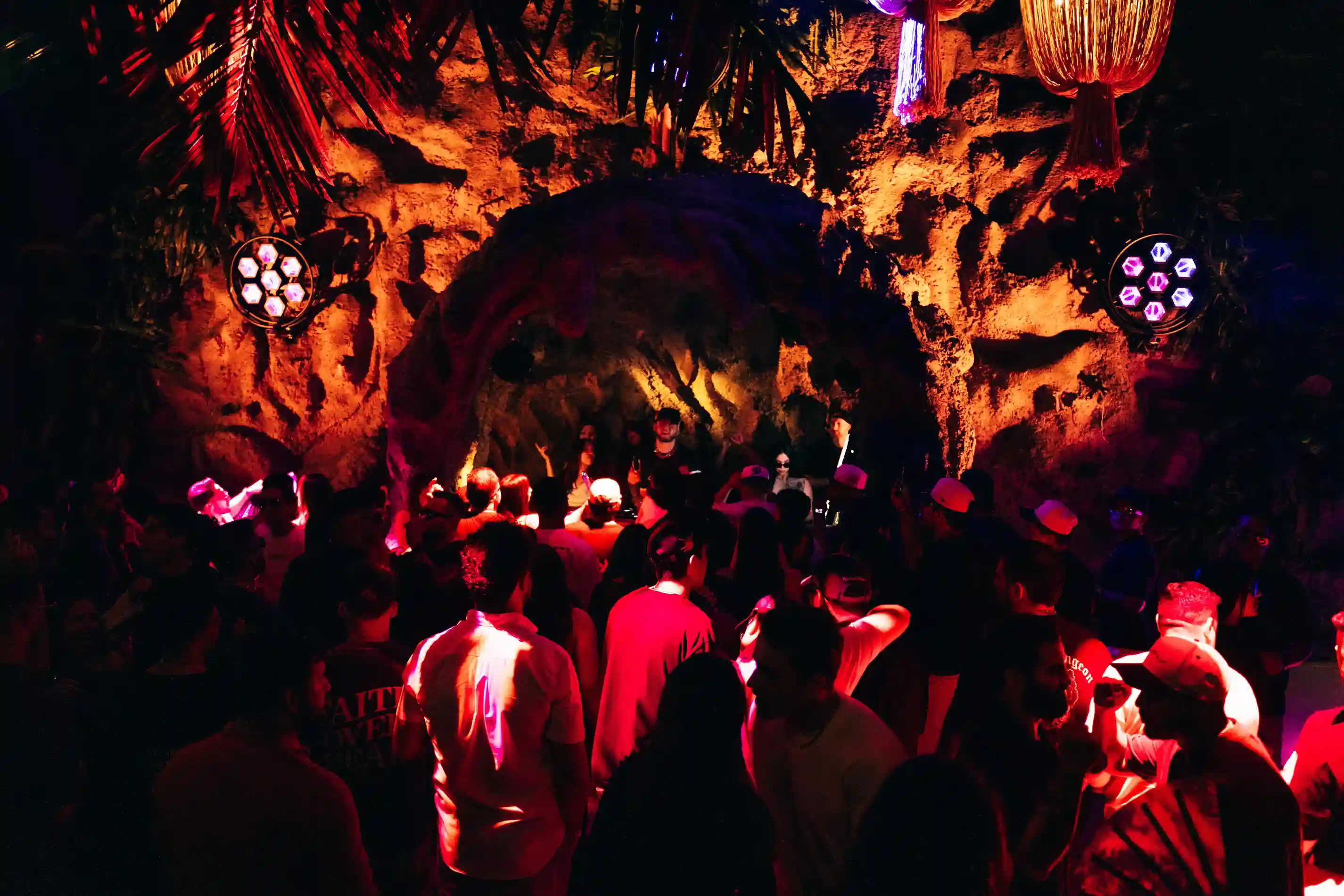 Crowd of people enjoying a dimly lit club with red and purple lights near a textured cave-like wall.