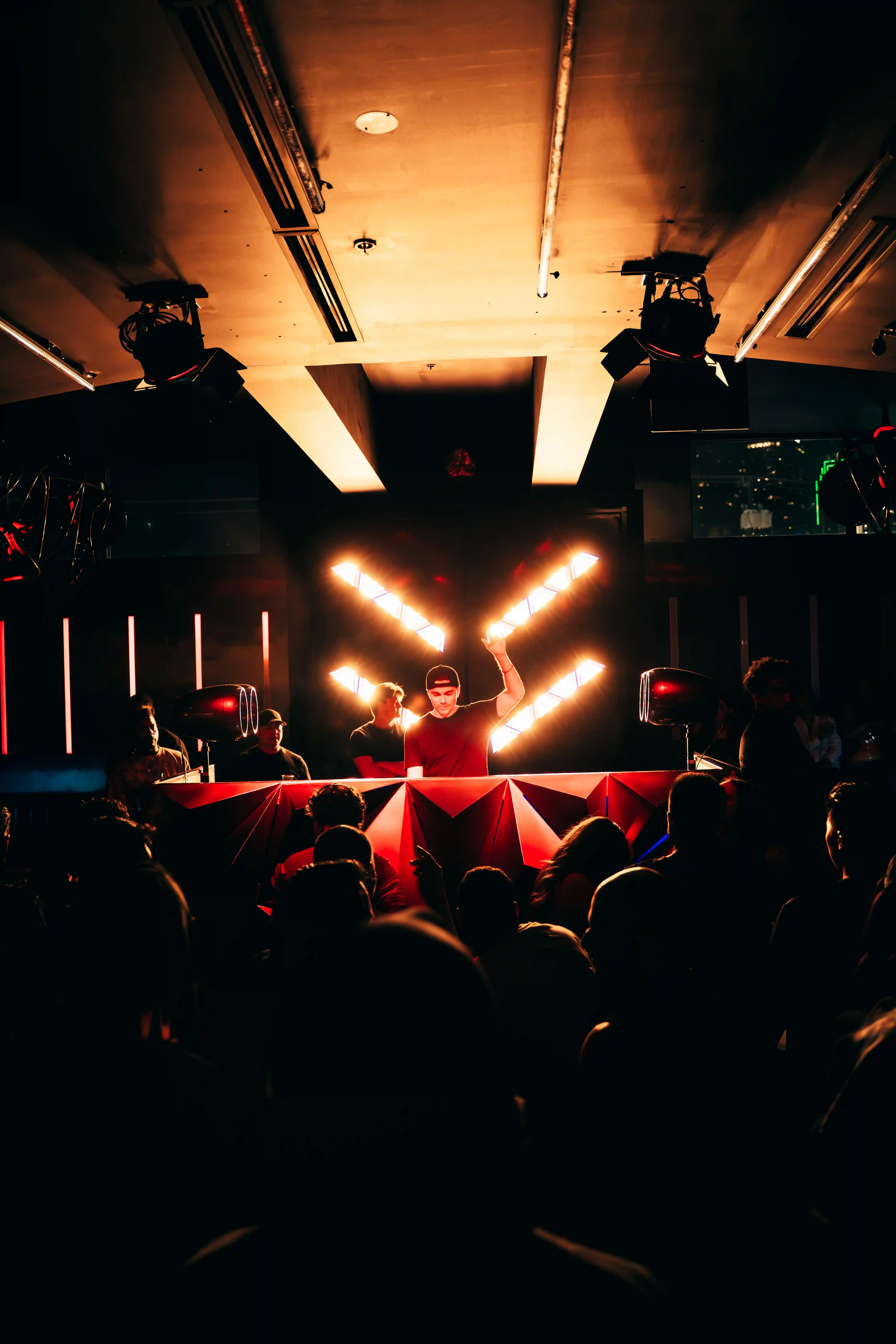 DJ performing at a nightclub with bright lights behind and a crowd in front.