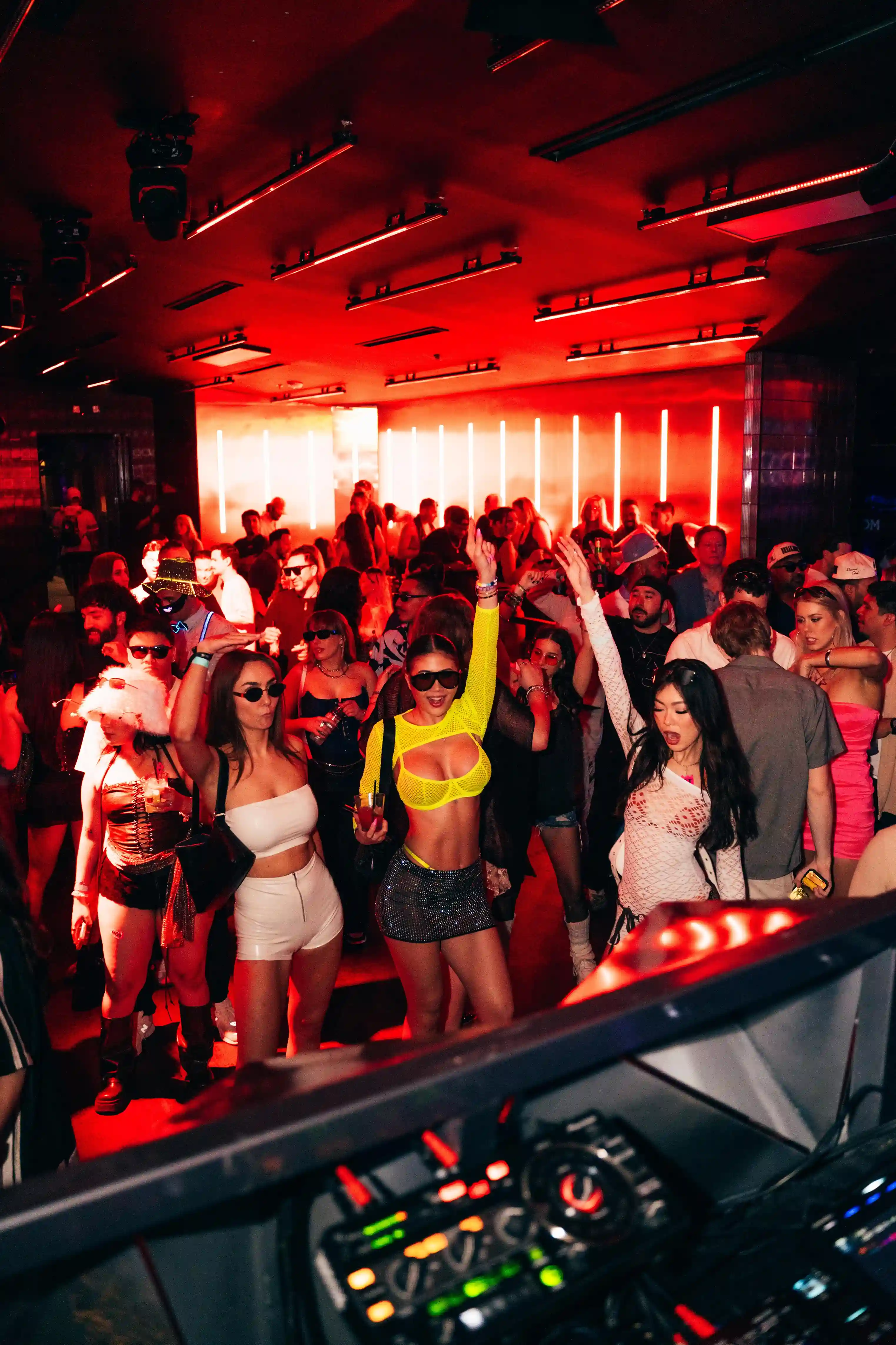 Crowd dancing in a nightclub with red lighting and a DJ mixer in the foreground.