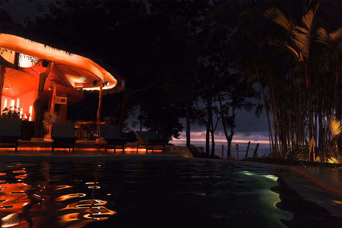 Night view of a poolside with lit lounge chairs and a thatched-roof structure, trees silhouetted against a twilight sky over the water.