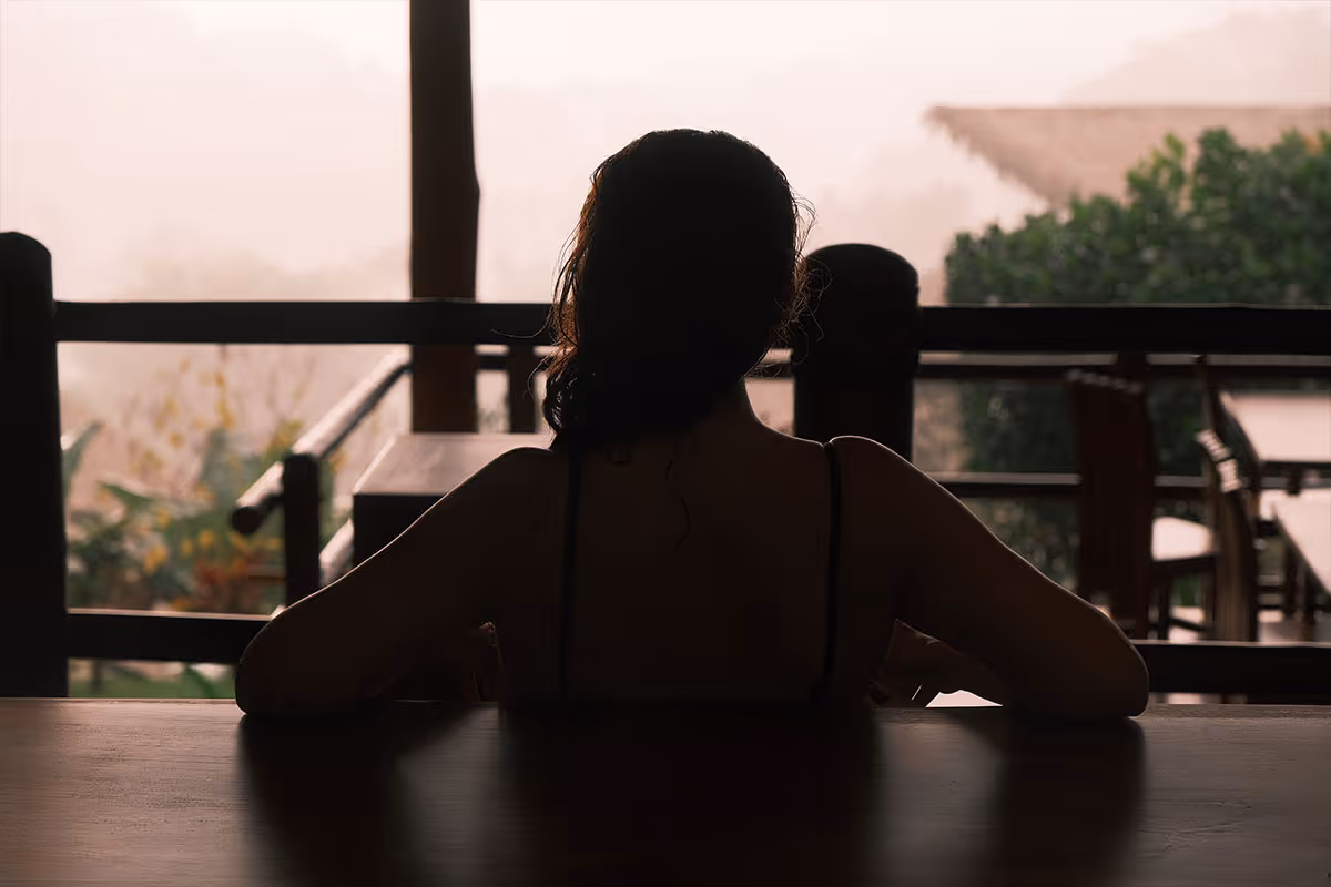 Silhouette of a woman sitting at a wooden table on a porch overlooking misty greenery.