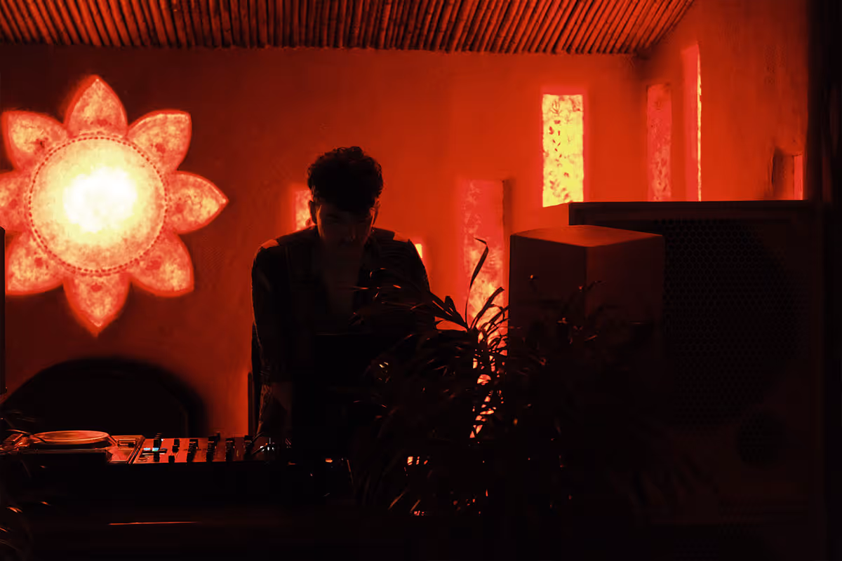 Person working at a DJ mixer in a dimly lit room with warm red lighting and a glowing flower-shaped wall decoration.