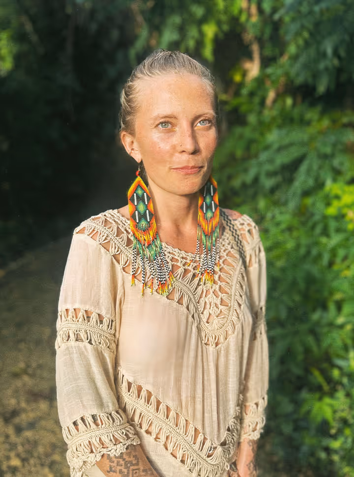 Woman with blonde hair tied back wearing large colorful beaded earrings and a beige crocheted top, standing outdoors in front of green foliage.