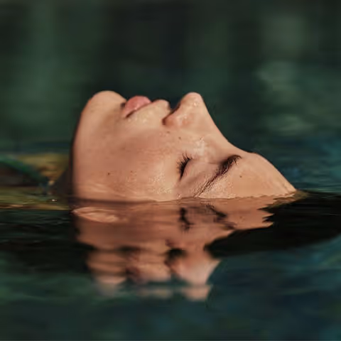 Close-up of a person's face with closed eyes partially submerged and reflected in calm water.