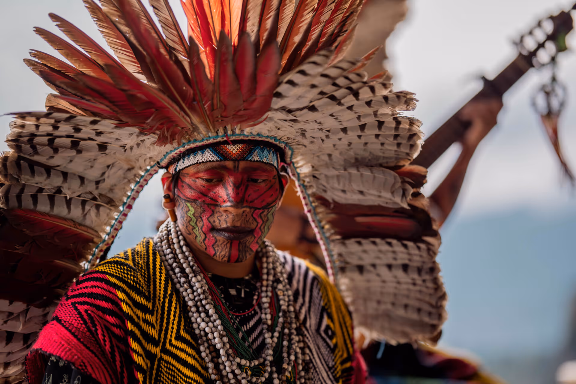 Person with red and black face paint wearing a large feathered headdress and colorful patterned clothing with layered white beaded necklaces.