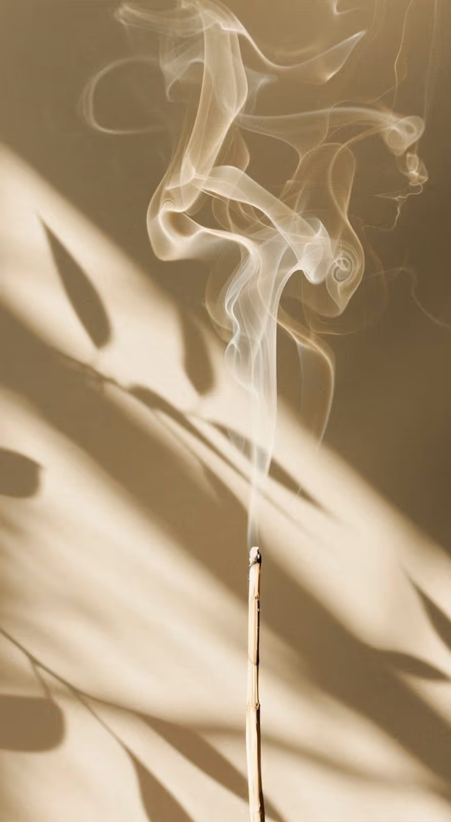 Thin incense stick burning with delicate tendrils of smoke curling upward against a beige background with leaf shadows.